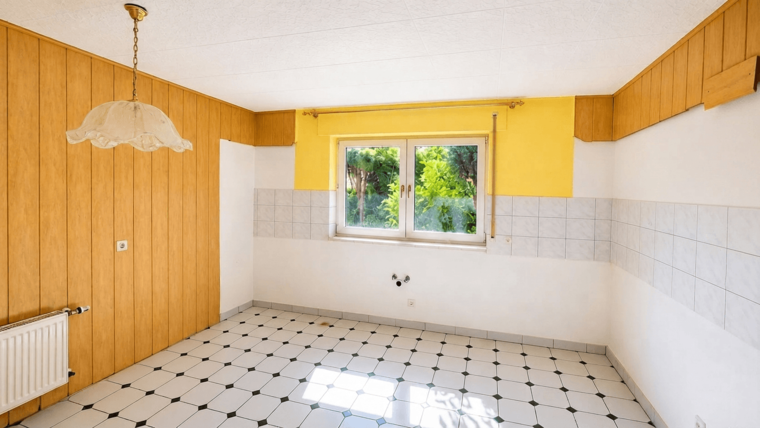 Ein heller, leerer Raum mit einer großen Fensteröffnung, die Blick auf grüne Bäume bietet. Die Wände sind teilweise in Holzoptik und gelb gestrichen. Der Boden besteht aus weißen Fliesen mit schwarzen Akzenten. Eine auffällige, hängende Lampe ist an der Decke angebracht.