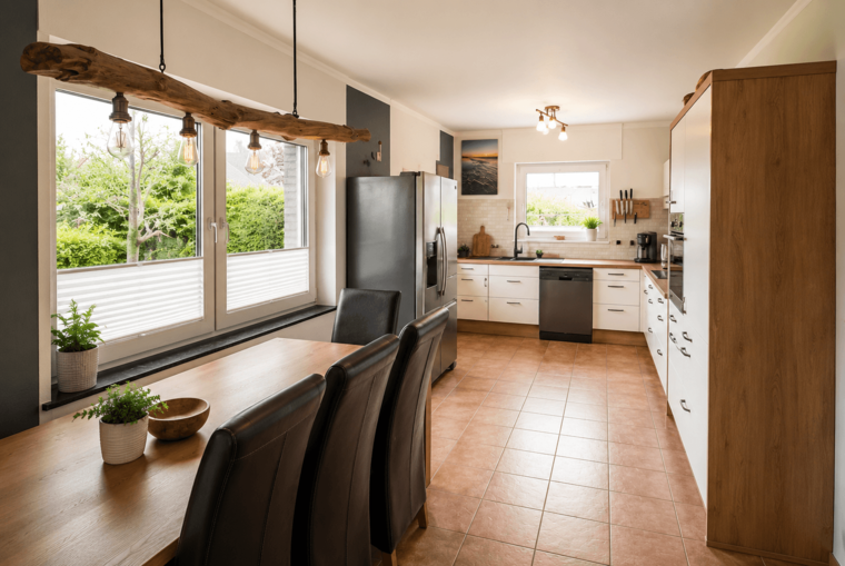Eine moderne Küche mit einem Holztisch und schwarzen Stühlen. Ein Kühlschrank sowie eine Spüle und ein Geschirrspüler sind sichtbar. Große Fenster lassen viel Tageslicht herein und bieten einen Blick auf grüne Pflanzen im Freien. Die Wände sind in hellen Farben gehalten.