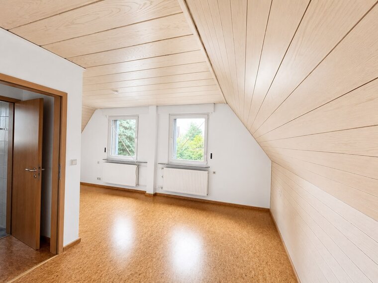 Ein heller, leerer Raum mit schräger Decke und Holzvertäfelung. Es gibt zwei Fenster, die Tageslicht hereinlassen, sowie einen Holzfußboden. Eine Tür führt zu einem angrenzenden Raum, der teilweise sichtbar ist. Der Raum wirkt geräumig und einladend.