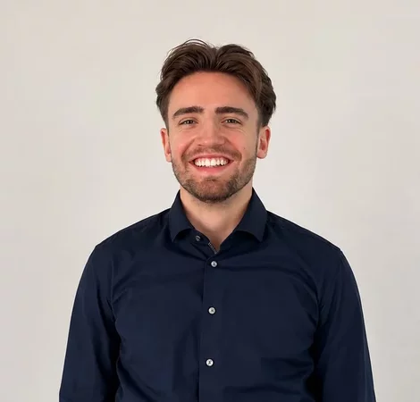 Ein junger Mann mit kurzen, braunen Haaren und einem gepflegten Bart lächelt in die Kamera. Er trägt ein dunkelblaues Hemd und steht vor einem neutralen, hellen Hintergrund. Sein Ausdruck wirkt freundlich und einladend.