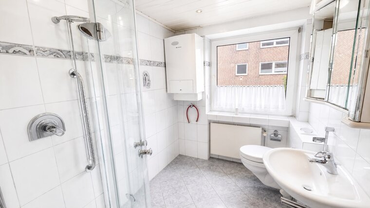 Das Bild zeigt ein modernes Badezimmer mit einem Glaskabine für die Dusche, einem Waschbecken und einer Toilette. Weiße Fliesen und eine große Fensterfront sorgen für helles Licht. Ein Heizkörper und ein Spiegel sind ebenfalls sichtbar.