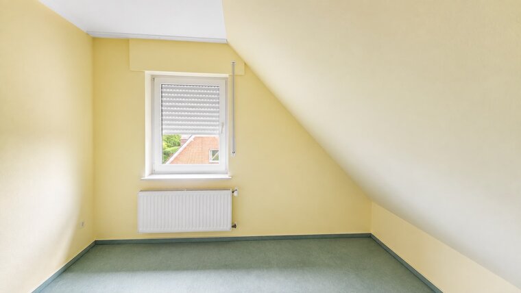 Ein heller Raum mit gelben Wänden und einem schrägen Dach. An einer Wand befindet sich ein Fenster mit Jalousien, durch das man einen Teil eines Gartens sieht. Der Boden ist in einem sanften Grünton gehalten.