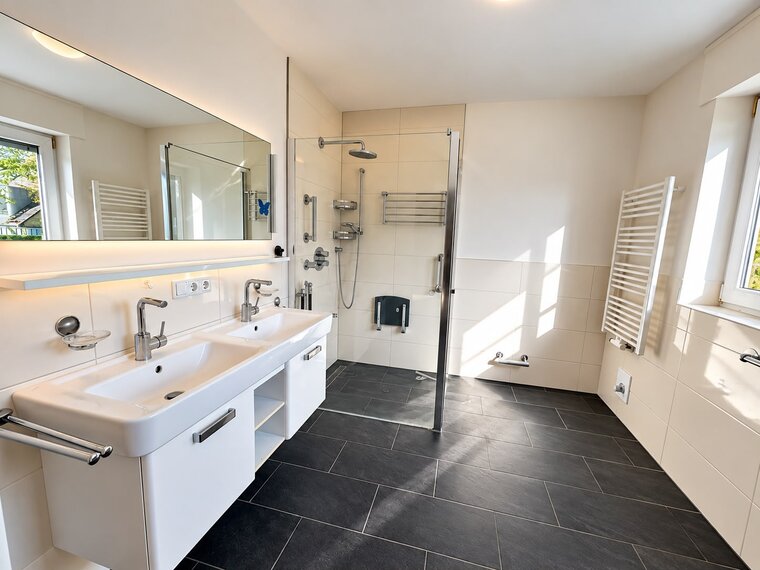 Das Bild zeigt ein modernes Badezimmer mit einem Doppelwaschbecken und Wasserhähnen. Es gibt eine große Dusche mit transparenter Glastür und verschiedene Duscharmaturen. Die Wände sind hell, der Boden ist dunkel gefliest, und das Zimmer hat Fenster, die Tageslicht hereinlassen.