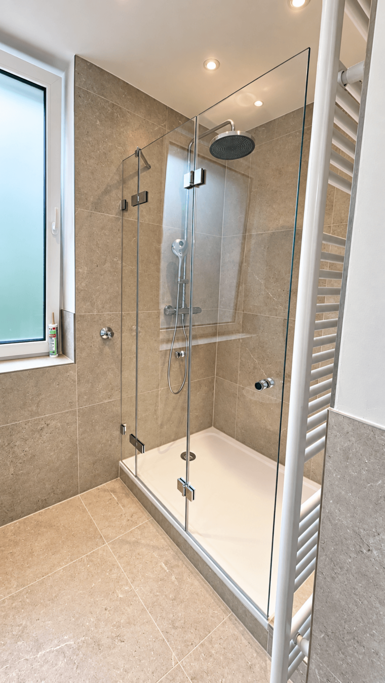 Ein modernes Badezimmer mit einer gläsernen Dusche. Die Wände sind in grauem Stein gehalten, und der Duschkopf hängt an der Wand. Neben der Dusche befindet sich ein Fenster, das Tageslicht hereinlässt, und ein Handtuchhalter aus weißem Metall.