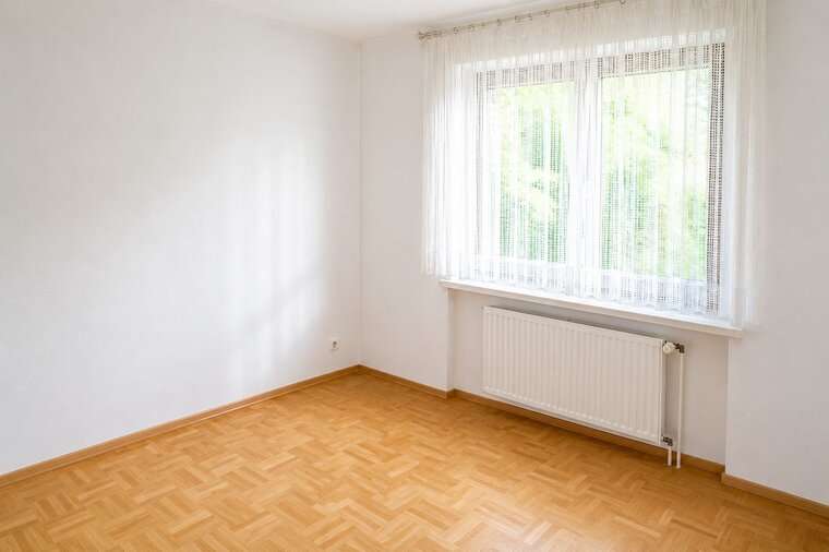 Ein leerer Raum mit hellen Wänden und einem großen Fenster, das Tageslicht hereinlässt. Der Fußboden ist aus hellbraunen Holzdielen. Vor dem Fenster hängt ein transparenter Vorhang, und an der Wand befindet sich eine Heizungsanlage.