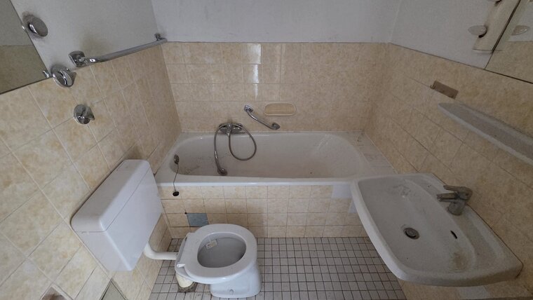 Das Bild zeigt ein Badezimmer mit einer Badewanne, einem Waschbecken und einer Toilette. Die Wände sind hell gefliest, und es gibt einen Spiegel. Die Badewanne enthält einen Wasserhahn und eine Handbrause. Das Gesamtbild wirkt schlicht und unauffällig.