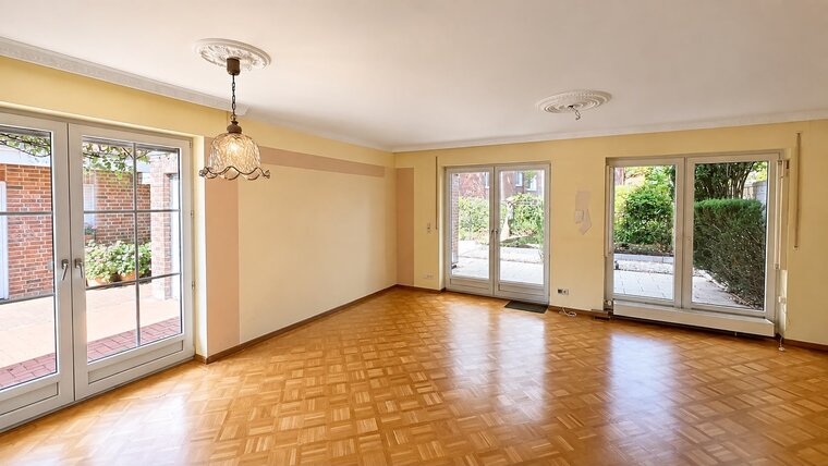 Ein helles Wohnzimmer mit parkettartigem Boden und großen Fenstern, die auf einen Außenbereich mit Pflanzen blicken. Eine dekorative Deckenlampe hängt in der Mitte des Raumes, der in hellen Farben gestrichen ist.