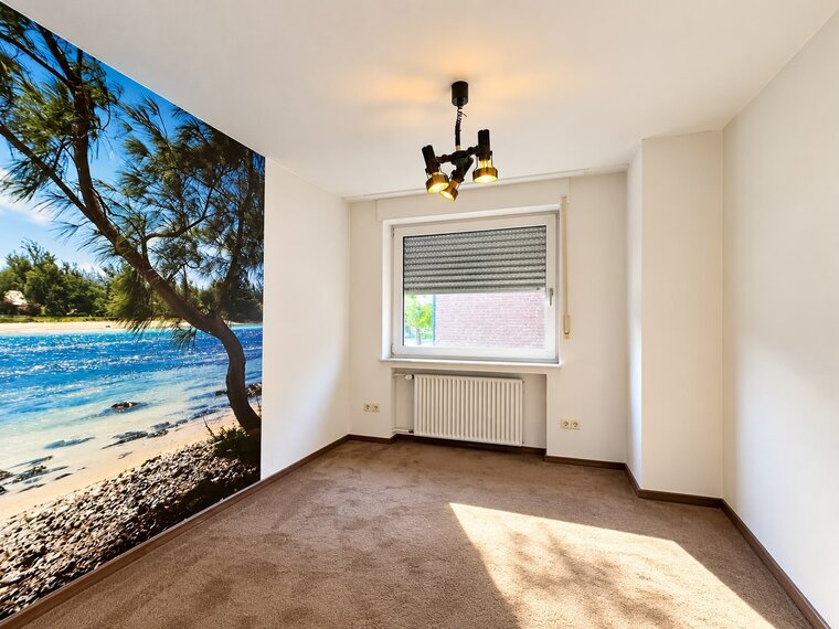 Ein helles Zimmer mit einem großen Fenster und einem Boden aus braunem Teppich. Eine Wand ist mit einem Bild eines Strandes dekoriert, das einen klaren blauen Himmel, Wasser und einen Baum zeigt. Die Beleuchtung wird durch eine schwarze Deckenlampe bereitgestellt.