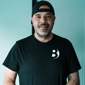 Ein Mann mit einem kurzen Bart trägt ein schwarzes T-Shirt mit einem weißen Logo. Er steht vor einer hellen Wand und lächelt. Der Mann hat eine Kappe auf und wirkt freundlich und zugänglich.