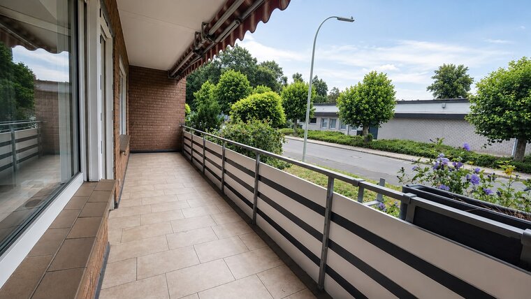 Ein geräumiger Balkon mit Fliesenboden, versehen mit einer Metallgeländer und einer Aussicht auf grüne Bäume und Sträucher. Im Hintergrund sind Wohnungen und eine Straße sichtbar. Der Himmel ist blau mit einigen Wolken.