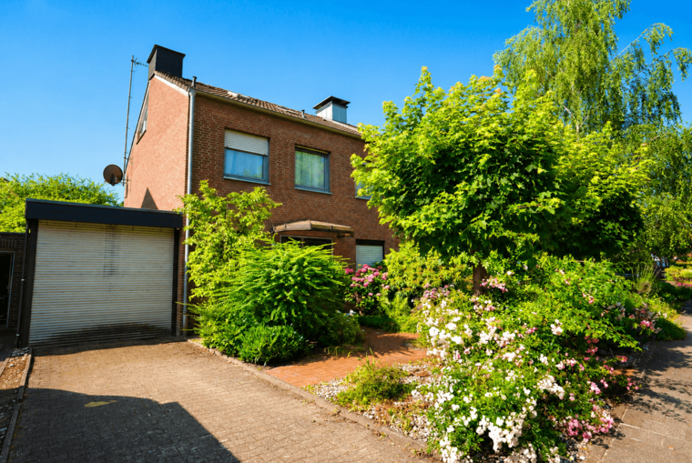 Ein zweigeschossiges, ziegelfarbenes Haus mit grünen Sträuchern und bunten Blumen im Vordergrund. Der Himmel ist blau und klar. Eine Einfahrt führt zum Garagentor, der Garten ist gepflegt und bietet eine lebendige, einladende Atmosphäre.
