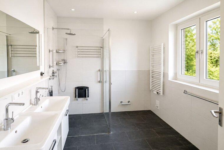 Das Bild zeigt ein modernes Badezimmer mit zwei Waschbecken, einer gläsernen Dusche und einem großen Spiegel. Es gibt helles Licht, helle Wände und einen grauen Fliesenboden. Ein Fenster bietet Blick ins Grüne. Die Einrichtung ist praktisch und zeitgemäß.