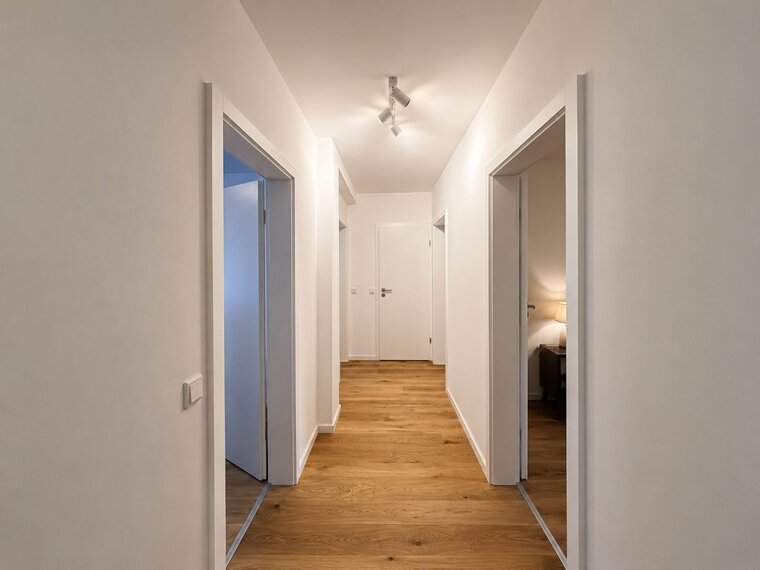 Ein heller Flur mit mehreren Türen auf beiden Seiten. Der Boden ist aus Holz, und die Wände sind in neutralen Tönen gehalten. Am Ende des Flurs befindet sich eine offene Tür, durch die sanftes Licht strahlt.