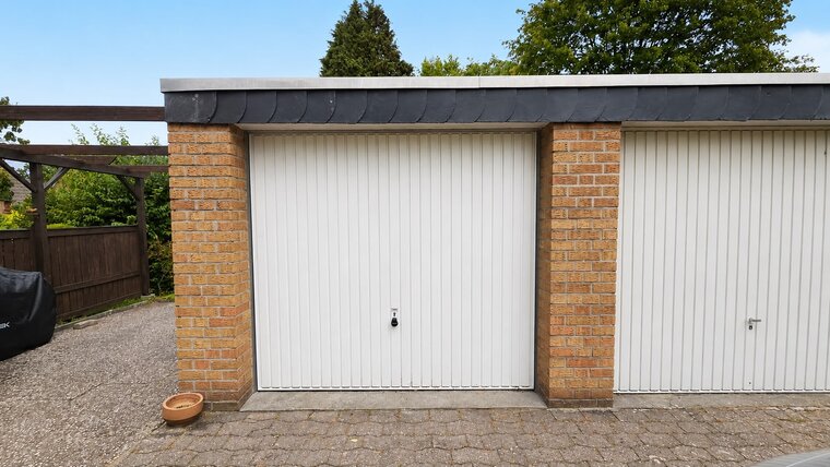 Eine Garage mit einem weißen Rolltor aus vertikalen Lamellen. Die Wände sind aus braunen Ziegelsteinen. An der linken Seite ist ein kleiner blumenförmiger Blumentopf sichtbar. Im Hintergrund sind Bäume und ein blauer Himmel zu erkennen.