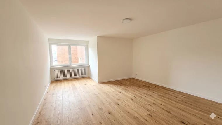 Ein heller, leerer Raum mit Holzfußboden. An einer Wand befindet sich ein großes Fenster, das Tageslicht hereinlässt. Auf der gegenüberliegenden Seite gibt es eine Heizung. Die Wände sind schlicht und neutral gehalten. Der Raum wirkt freundlich und einladend.