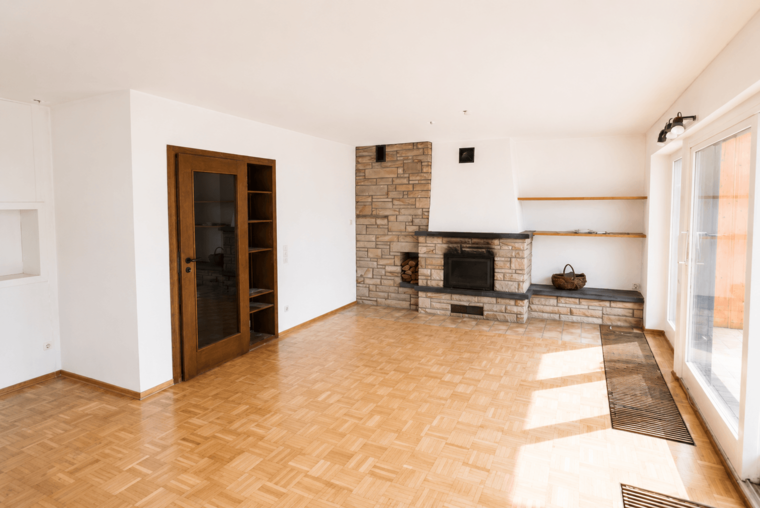 Ein heller Wohnraum mit Holzfußboden und einem modernen Kamin aus Stein. An einer Wand befindet sich ein Regal sowie eine gläserne Tür, die zu einem Außenbereich führt. Der Raum ist lichtdurchflutet und bietet viel Platz.