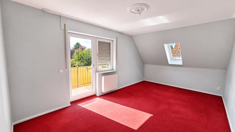 Ein heller Raum mit rotigem Teppichboden. Eine Fenstertür öffnet sich zu einem Balkon, durch das Fenster ist ein Teil des Außenbereichs mit Bäumen zu sehen. Die Wände sind hellblau, und es gibt eine Heizung an der Wand.