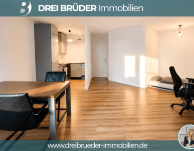 Ein helles, modernes Zimmer mit Holzfußboden. Auf der linken Seite befindet sich eine Küchenzeile mit Schränken und einem Esstisch. Rechts steht ein Schreibtisch mit einem Stuhl. An der Wand ist ein Licht angebracht, und Sonnenschein fällt durch ein Fenster.