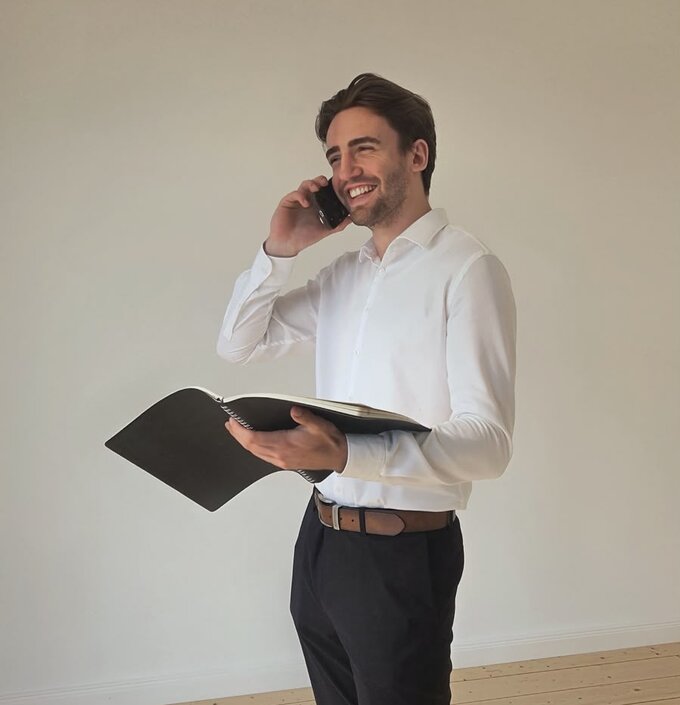 Ein lächelnder Mann steht in einem hellen Raum und spricht telefonisch. Er trägt ein weißes Hemd und schwarze Hose und hält eine schwarze Akte in der Hand. Der Boden ist aus hellem Holz.