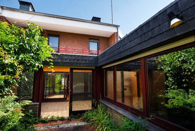 Ein rechteckiger Innenhof ist von einem modernen Gebäude mit Holz- und Glaswänden umgeben. Die Pflanzen auf dem Boden und die grünen Kletterpflanzen sorgen für eine harmonische Verbindung zur Natur. Das Dach ist mit dunklen Schindeln bedeckt, und der Himmel ist klar und blau.