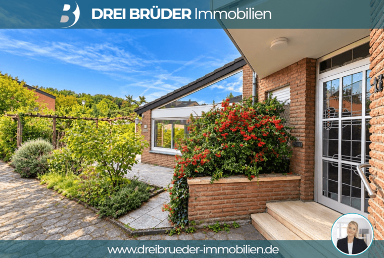 Eine moderne, einladende Hausansicht mit einem gepflegten Garten und bunten Blüten. Der Eingangsbereich ist gut beleuchtet, und die Fassade ist aus roten Ziegeln. Im Hintergrund sind grüne Bäume und Sträucher sichtbar, die eine freundliche Atmosphäre schaffen.