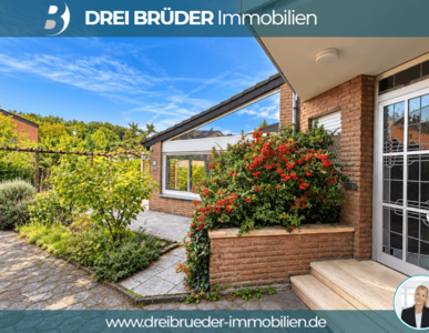 Eine moderne, einladende Hausansicht mit einem gepflegten Garten und bunten Blüten. Der Eingangsbereich ist gut beleuchtet, und die Fassade ist aus roten Ziegeln. Im Hintergrund sind grüne Bäume und Sträucher sichtbar, die eine freundliche Atmosphäre schaffen.