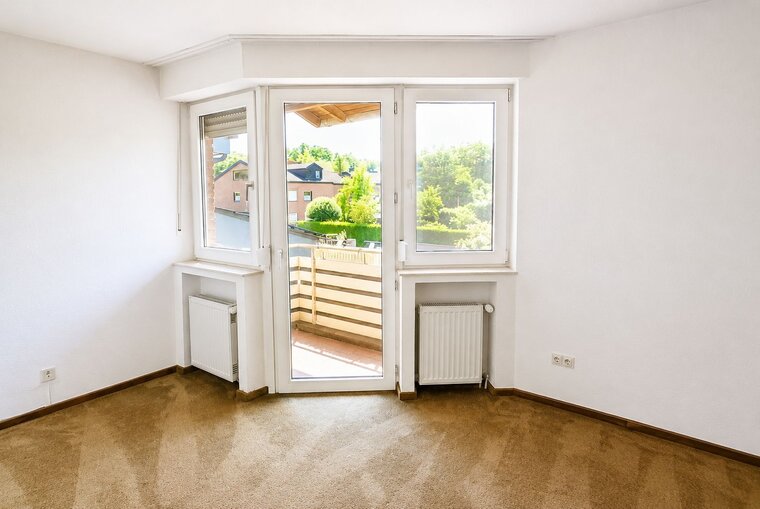 Ein lichtdurchfluteter Raum mit Teppichboden. An einer Wand befindet sich ein Fenster mit Zugang zu einem Balkon, der den Blick auf eine grüne Landschaft und benachbarte Häuser freigibt. Heizkörper sind an den Wänden installiert. Der Raum hat eine helle, neutrale Farbgebung.