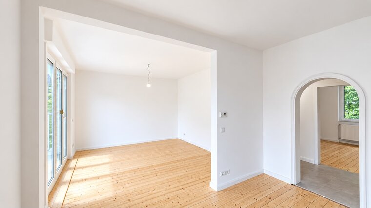 Innenansicht eines hellen Raumes mit Holzfußboden, weißen Wänden und großen Fenstern. Ein einfacher Lampenanschluss hängt von der Decke. Im Hintergrund ist ein Durchgang zu einem weiteren Raum sichtbar, der ebenfalls hell erleuchtet ist.