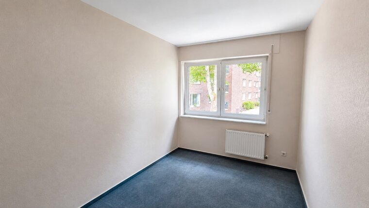 Ein heller, leerer Raum mit beige gestrichenen Wänden und einem blauen Teppichboden. An einer Wand befindet sich ein großes Fenster, durch das Licht eintritt und einen Blick auf einen Baum und ein weiteres Gebäude zeigt. Heizung unter dem Fenster.
