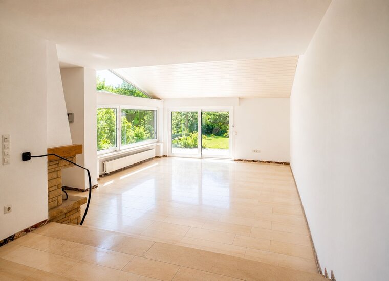 Ein helles, geräumiges Wohnzimmer mit großen Fenstern, die auf einen grünen Garten blicken. Die Wände sind weiß und der Boden ist mit hellen Fliesen ausgelegt. Ein Kamin ist in einer Ecke sichtbar, und die Decke hat eine geneigte Form.