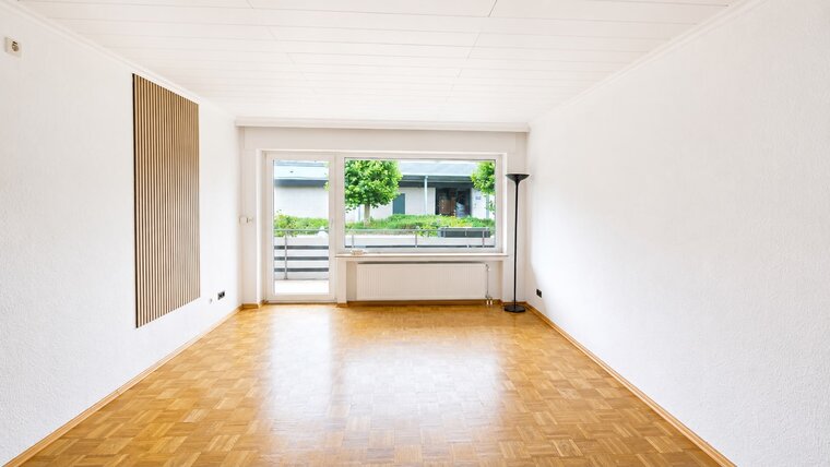 Ein heller, leerer Raum mit Parkettboden und weißen Wänden. Große Fenster lassen viel Tageslicht herein und bieten einen Blick auf einen Balkon und Pflanzen im Freien. An der Wand steht eine Stehlampe. Der Raum wirkt einladend und geräumig.
