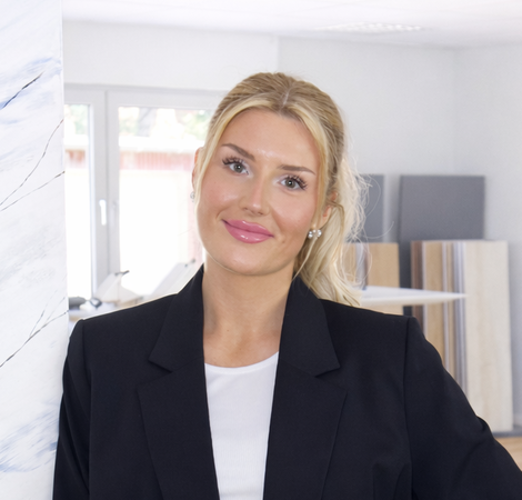 Eine junge Frau steht an einer Wand mit einer marbled Textur. Sie trägt ein schwarzes Blazer-Outfit und lächelt. Im Hintergrund sind Materialien wie Holzplatten sichtbar, was auf ein kreatives oder geschäftliches Umfeld hindeutet.