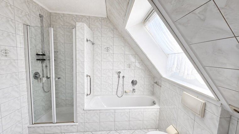 Das Bild zeigt ein modernes Badezimmer mit einer schrägen Decke. Auf der linken Seite befindet sich eine Duschkabine mit Glastüren, rechts eine Badewanne. Die Wände sind mit hellen Fliesen gefliest, und ein Dachfenster lässt Tageslicht herein.