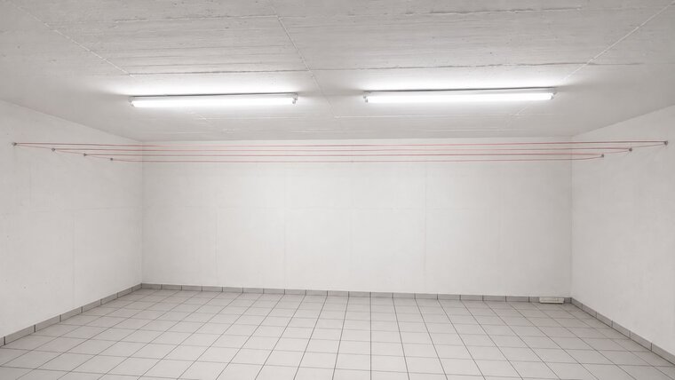 Ein leeres, weißes Zimmer mit einem Fliesenboden. An der Wand sind einige rote Linien angebracht, die horizontal verlaufen. Die Decke ist beleuchtet mit zwei langen Leuchten, die eine helle Umgebung schaffen. Der Raum wirkt minimalistisch und offen.