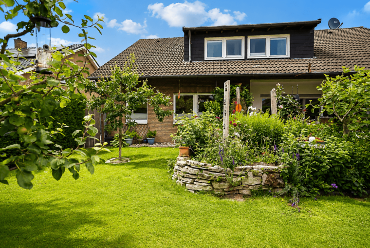 Ein modernes Einfamilienhaus mit einem großen Garten, der von üppigem Grün und bunten Blumen umgeben ist. Im Vordergrund steht ein steinerner Pflanzring mit Pflanzen. Der Himmel ist blau mit einigen Wolken.