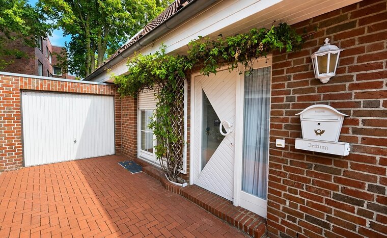 Eingangsbereich eines Hauses mit einer weißen Haustür, einem stilvollen Briefkasten mit der Aufschrift „Zeitung“ und einer Pflanze, die an der Wand wächst. Neben der Tür befindet sich eine Veranda aus roten Klinkersteinen und eine geschlossene Garage. Bäume sind im Hintergrund sichtbar.