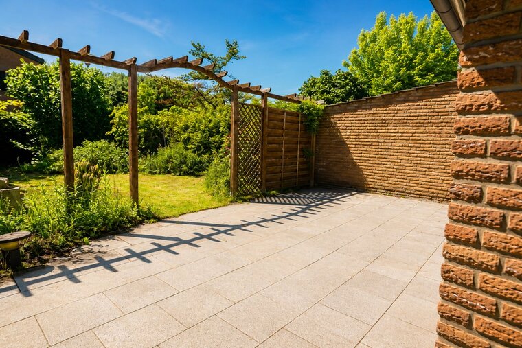 Ein sonniger Gartenbereich mit einem offenen Platz aus grauen Fliesen. An der Seite steht ein Holzpergola mit Pflanzen. Im Hintergrund sind hohe, grünende Sträucher und Bäume sichtbar, die eine ruhige, natürliche Atmosphäre schaffen.