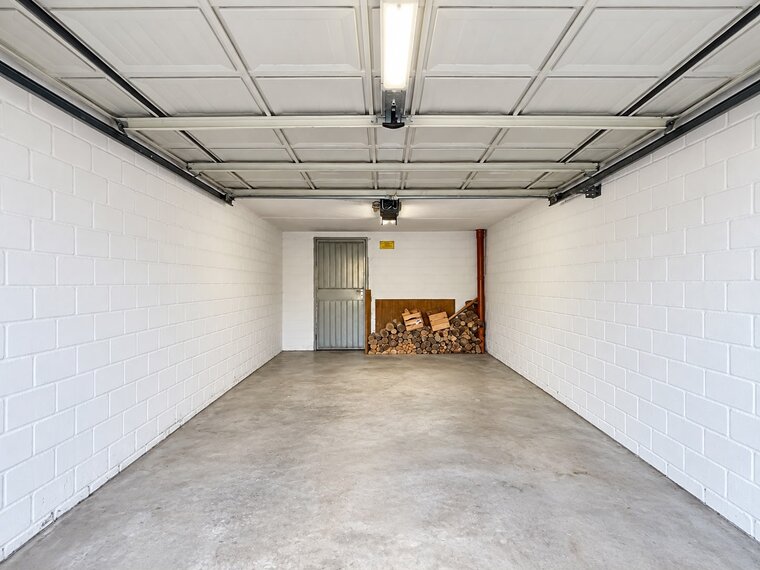 Ein leerer Garagenraum mit weißen Wänden und einem Betonboden. In einer Ecke sind gestapelte Holzscheite auf einem Holzbrett zu sehen. Ein Metalltor führt zu einem anderen Raum. Die Decke ist mit einer Beleuchtung ausgestattet.