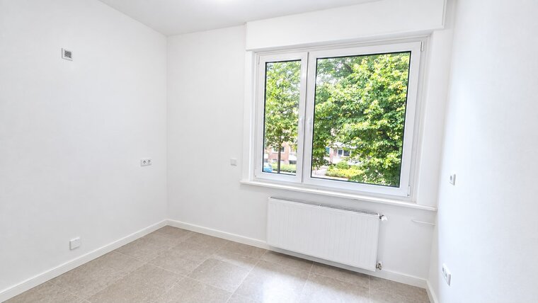 Ein heller, leerer Raum mit weißen Wänden und einem großen Fenster, durch das viel Tageslicht strömt. Draußen sind grüne Bäume sichtbar. Am Boden ist ein heller Fliesenboden zu sehen, und an der Wand befindet sich eine Heizungsanlage.