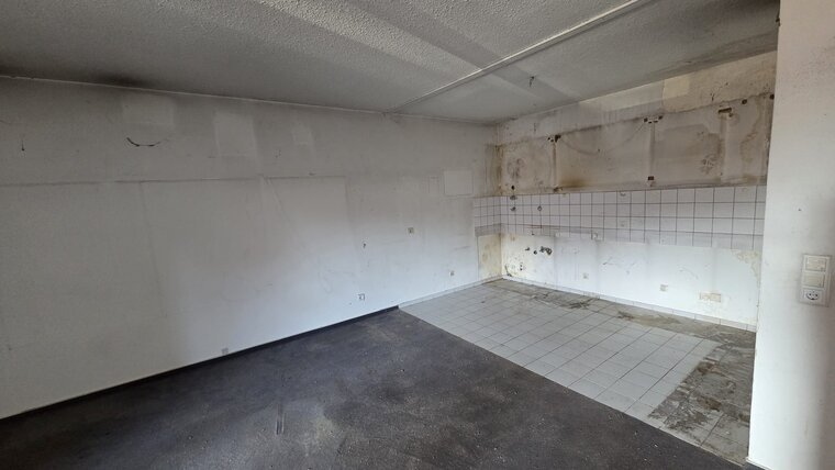 Ein leerer Raum mit einer gefliesten Wand. Auf dem Boden liegt dunkler Teppichboden, während die Wand Flecken und Schäden aufweist. Die Fliesen in der Ecke sind teilweise schmutzig, und die Wände sind fast vollständig weiß, zeigen aber Abnutzungsspuren.