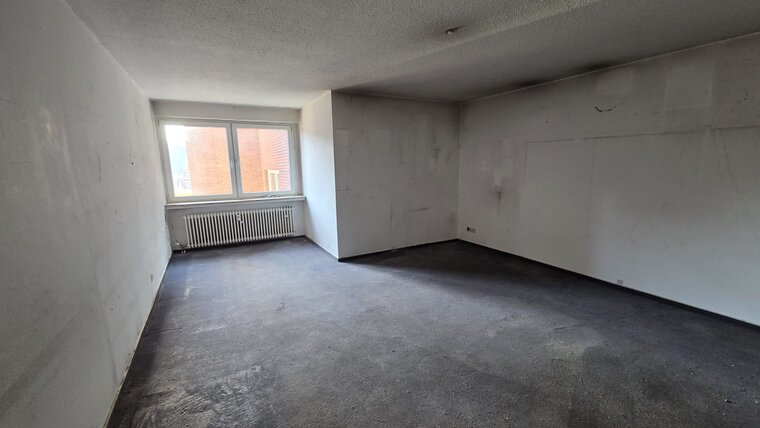 Ein leerer Raum mit weißen Wänden und einem Fenster, das Tageslicht hereinlässt. Der Fußboden ist grau und unbeleuchtet. An der Wand unter dem Fenster befindet sich ein Heizkörper. Der Raum wirkt unmöbliert und neutral.