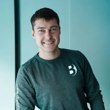Ein junger Mann mit braunen Haaren lächelt freundlich in die Kamera. Er trägt ein dunkelgrünes Sweatshirt mit einem weißen Logo. Der Hintergrund ist hell und einfarbig.