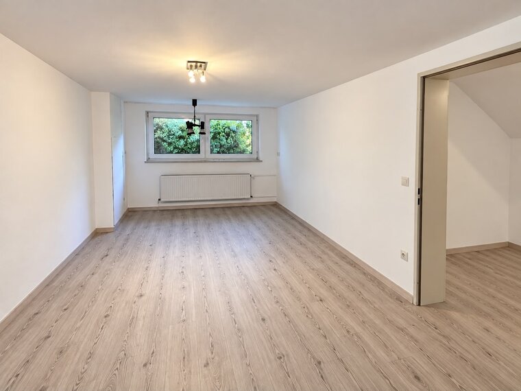Helle, leere Wohnräume mit Holzfußboden. Große Fenster lassen Tageslicht herein und bieten Blick auf grünes Laub. Eine Tür führt in einen angrenzenden Raum. Reliquien wie Heizkörper und Deckenbeleuchtung sind sichtbar, der Raum wirkt einladend und offen.