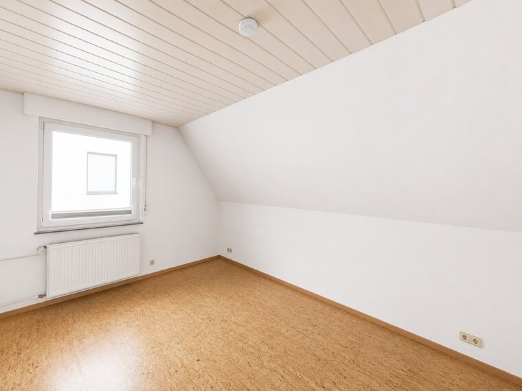 Ein leerer Raum mit schrägen Wänden und einem Fenster, das Tageslicht hereinlässt. Der Boden besteht aus hellem Holz oder Kork. Es gibt eine Heizkörper unter dem Fenster. Die Wände und Decke sind in neutralen Weißtönen gehalten.