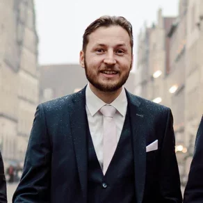 Ein lächelnder Mann in einem dunkelblauen Anzug mit hellrosa Krawatte steht in einer Stadtstraße. Leichte Nässe ist auf seinem Anzug sichtbar, während er in Richtung Kamera blickt. Im Hintergrund sind verschwommene Gebäude zu erkennen.