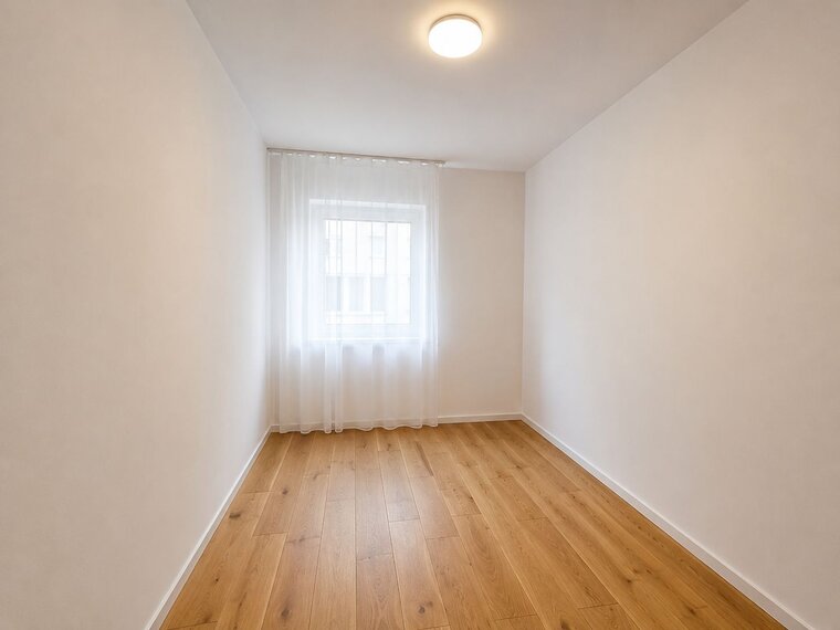 Ein helles, leeres Zimmer mit einem Fenster, das von einem weißen Vorhang umgeben ist. Der Boden ist aus Holz, und die Wände sind in neutralen Farben gehalten. Die Decke hat eine einfache Lampe, die das Zimmer beleuchtet.