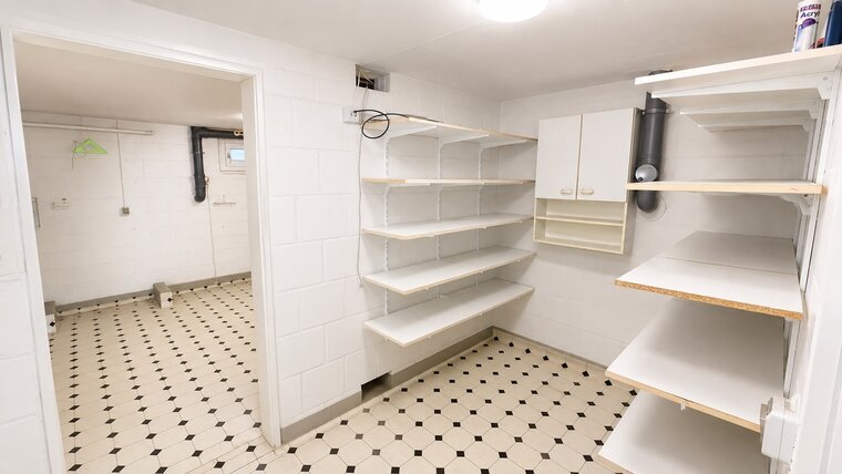 Ein heller Raum mit weißen Wänden und einem schwarzen und weißen Fliesenboden. An den Wänden befinden sich mehrere Regale ohne Inhalt sowie ein Schrank. Ein Fenster ist sichtbar, und es gibt einen Hängeständer.