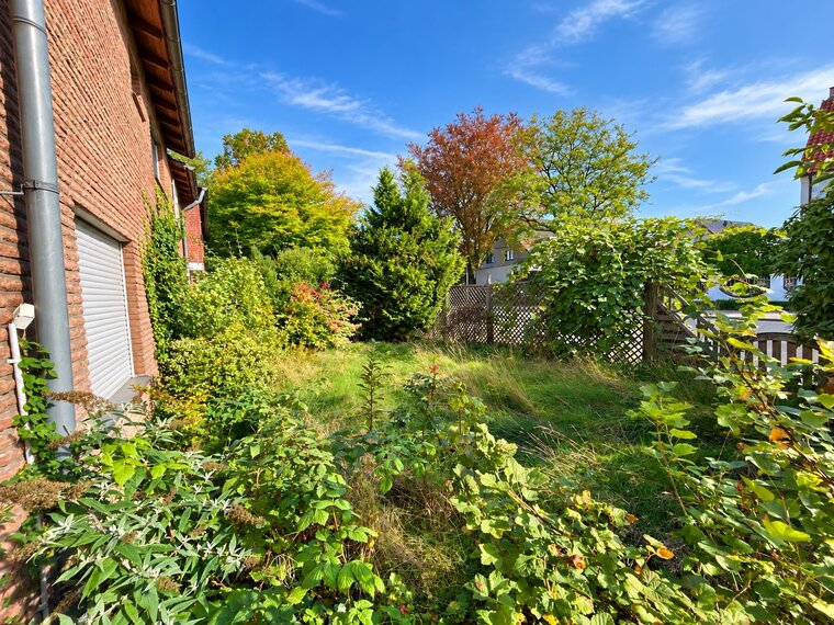 Eine grüne Gartenszene neben einem roten Ziegelhaus. Verschiedene Pflanzen und Bäume mit herbstlichen Farben sind sichtbar. Der Himmel ist hellblau mit einigen Wolken. Ein Zaun ist im Hintergrund zu erkennen. Das Gras ist unregelmäßig gewachsen und zeugt von wenig Pflege.