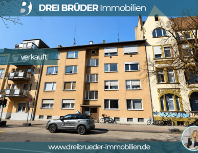 Das Bild zeigt ein mehrstöckiges Gebäude mit hellgelber Fassade. Ein Schild mit der Aufschrift „verkauft“ ist deutlich sichtbar. Auf der Straße parkt ein graues Auto, und im Hintergrund sind weitere Gebäude und Fahrräder zu sehen.