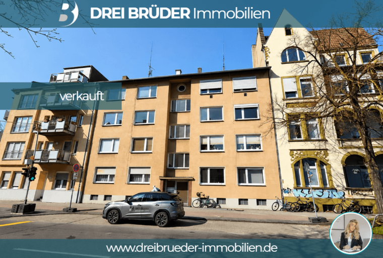 Das Bild zeigt ein mehrstöckiges Gebäude mit hellgelber Fassade. Ein Schild mit der Aufschrift „verkauft“ ist deutlich sichtbar. Auf der Straße parkt ein graues Auto, und im Hintergrund sind weitere Gebäude und Fahrräder zu sehen.